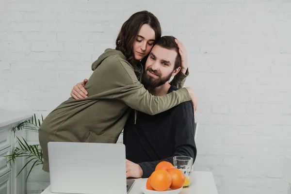 Hermosa pareja feliz abrazo mientras está sentado en la mesa con el ordenador portátil y naranjas durante el desayuno en la cocina - foto de stock