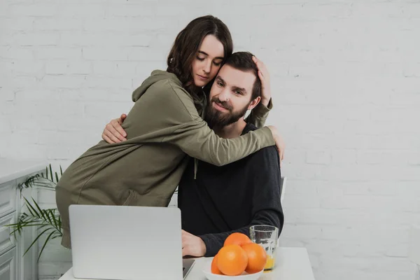 Beau couple heureux câlin tout en étant assis à la table avec ordinateur portable et oranges pendant le petit déjeuner dans la cuisine — Photo de stock