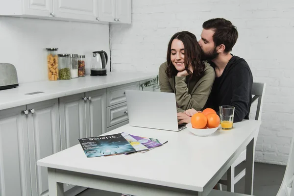 Handsome man hugging woman sitting at table with oranges and using laptop in kitchen — Stock Photo