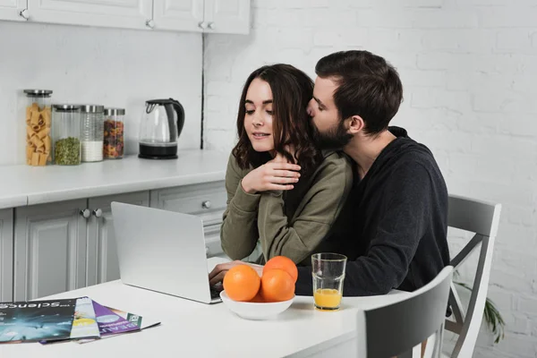 Homem bonito abraçando mulher sentada à mesa com laranjas e usando laptop na cozinha — Fotografia de Stock