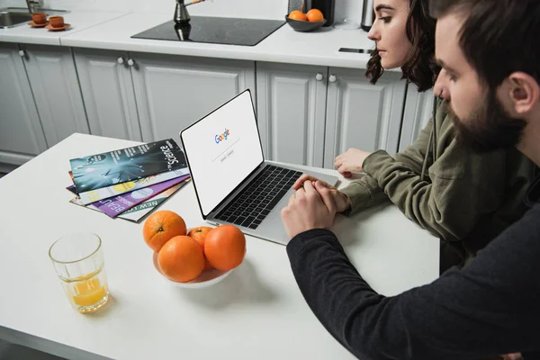 Couple sitting at table and using laptop with google website on screen in kitchen — Stock Photo