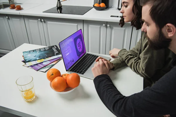 Casal sentado à mesa e usando laptop com site de compras on-line na tela na cozinha — Fotografia de Stock