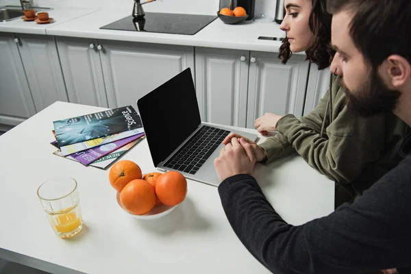 Pareja sentada en la mesa y utilizando el ordenador portátil con pantalla en blanco en la cocina - foto de stock