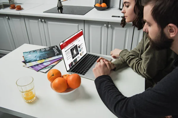 Pareja sentada en la mesa y el uso de ordenador portátil con BBC sitio web de noticias en la pantalla en la cocina - foto de stock