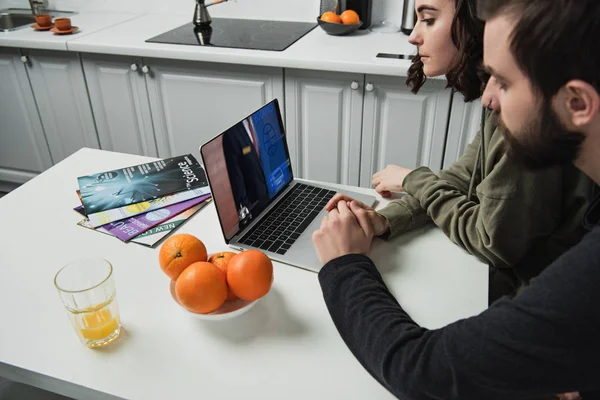 Casal sentado à mesa e usando laptop com site de reserva na tela na cozinha — Fotografia de Stock