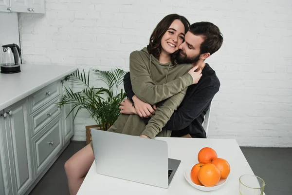 Schönes glückliches Paar, das sich beim Frühstück in der Küche am Tisch mit Laptop und Orangen umarmt — Stockfoto