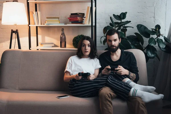 Beautiful surprised young couple sitting on couch with joysticks and playing video game in living room — Stock Photo