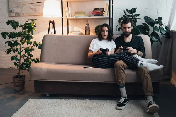 Schönes junges Paar sitzt auf Couch mit Steuerknüppeln und spielt Videospiel im Wohnzimmer mit Kopierraum — Stockfoto