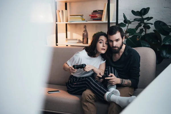 Hermosa pareja joven sentada en el sofá con joysticks y jugando videojuego en la sala de estar con espacio de copia - foto de stock