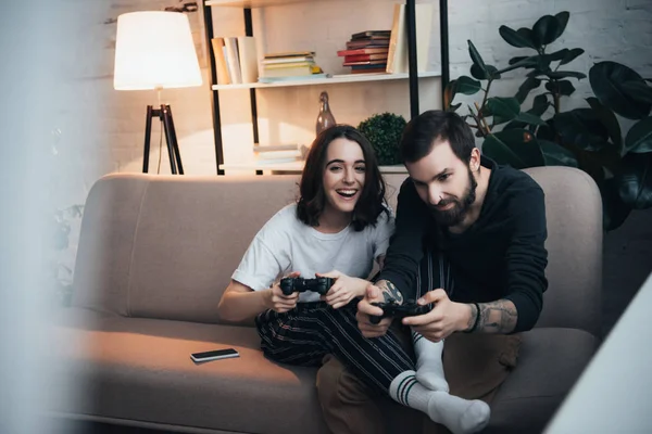 Селективное внимание красивой молодой пары, сидящей на диване с джойстиками и играющей в видеоигры в гостиной — стоковое фото