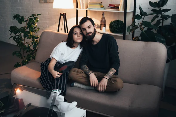 Hermosa joven pareja sentada en el sofá y mirando a la cámara mientras escucha el disco de vinilo en la sala de estar - foto de stock