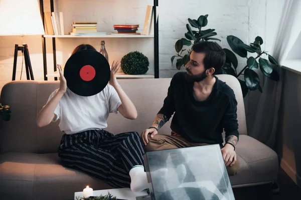 Mujer joven cubriendo la cara con disco de vinilo mientras está sentado en el sofá con el hombre en la sala de estar - foto de stock