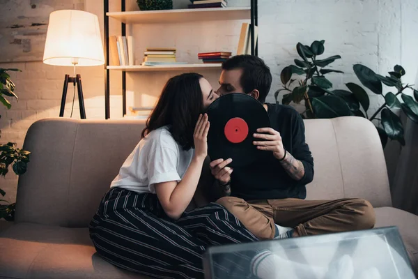 Hermosa pareja joven cubriendo caras con disco de vinilo y besándose mientras está sentado en el sofá en la sala de estar - foto de stock