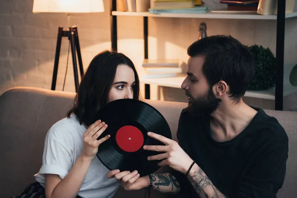 Hermosa mujer joven cubriendo la cara con disco de vinilo mientras está sentado en el sofá con el hombre en la sala de estar - foto de stock