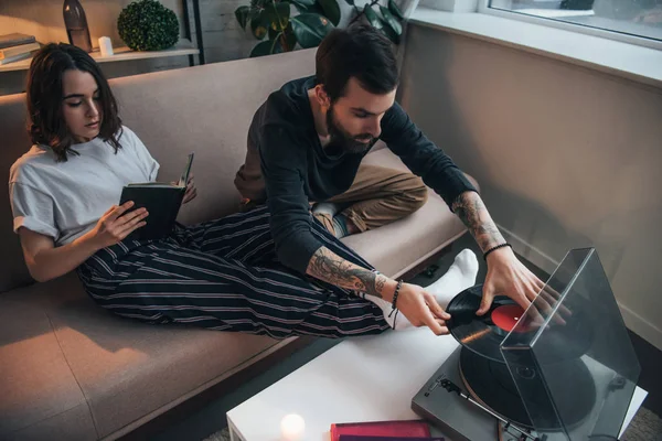 Hombre poniendo disco de vinilo en el tocadiscos, mientras que la mujer sentada en el sofá y libro de lectura en la sala de estar - foto de stock