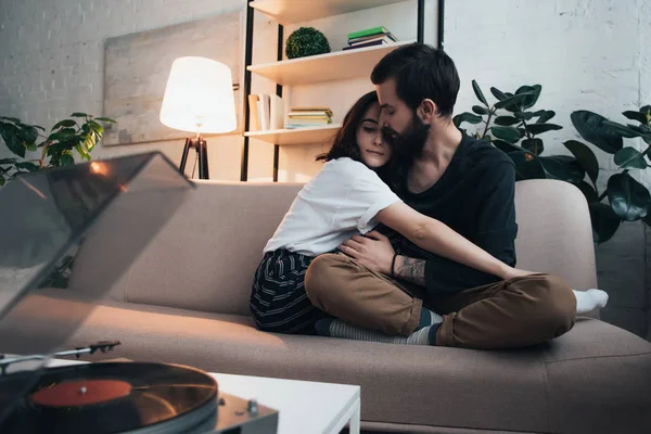 Beau jeune couple assis sur le canapé, étreignant et écoutant disque de vinyle dans le salon — Photo de stock