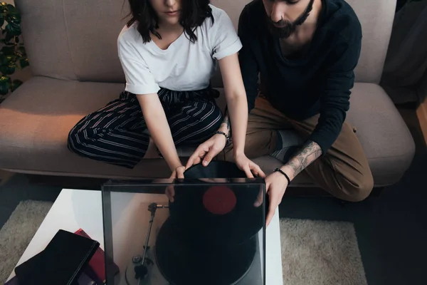 Vista parcial de pareja poniendo disco de vinilo en el tocadiscos mientras está sentado en el sofá - foto de stock