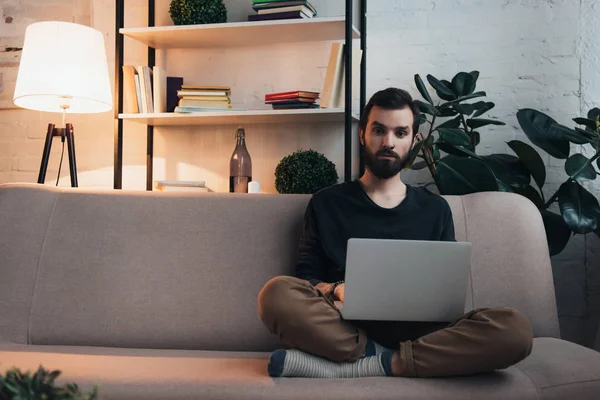 Schöner bärtiger Mann sitzt auf der Couch, schaut in die Kamera und benutzt Laptop im Wohnzimmer — Stockfoto