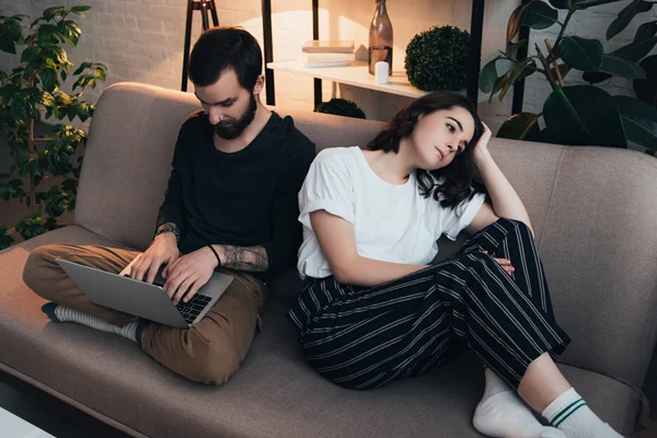 Mujer aburrida sentada en el sofá, mientras que el hombre usando el ordenador portátil en la sala de estar - foto de stock