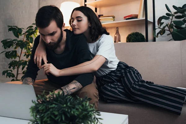 Mulher sentada no sofá e abraçando o homem usando laptop na sala de estar — Fotografia de Stock