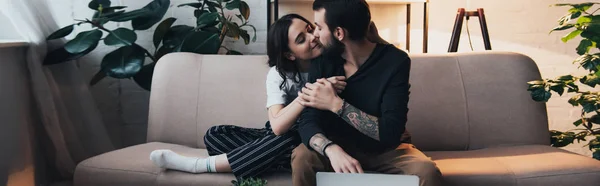 Hermosa joven tatuado pareja abrazando mientras sentado en sofá en sala de estar - foto de stock