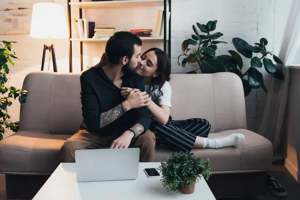 Hermosa pareja joven sentada en el sofá y besándose cerca de la computadora portátil y teléfono inteligente en la mesa en la sala de estar - foto de stock