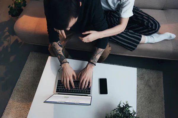 Cropped view of couple sitting ang hugging while using laptop with facebook website on screen at home — Stock Photo