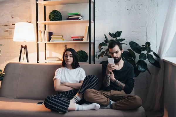 Gelangweilte Frau sitzt mit verschränkten Armen auf Couch, während Mann Smartphone im Wohnzimmer benutzt — Stockfoto