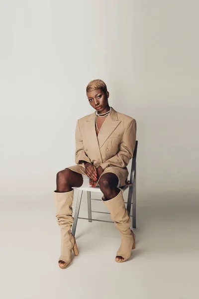 Mulher americana africana na moda em elegante jaqueta bege sentado na cadeira em cinza — Fotografia de Stock
