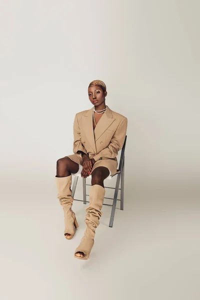 Elegante modelo afroamericano en chaqueta beige sentado en silla en gris - foto de stock