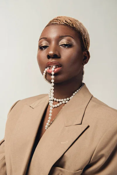 Портрет модной африканской женщины в бежевом пиджаке и ожерелье, изолированном на сером — стоковое фото