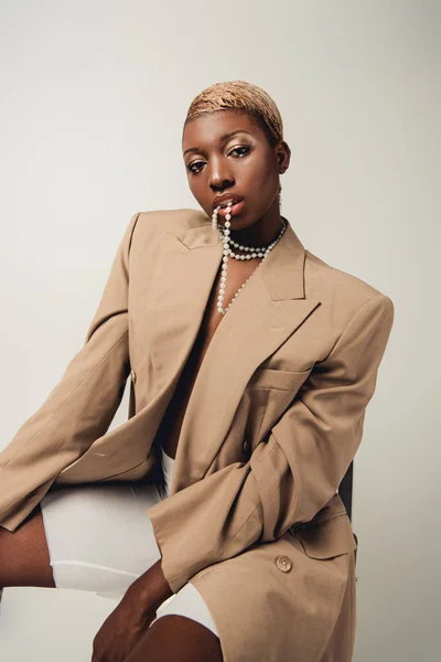 Donna afroamericana alla moda in collana e giacca beige seduta su sedia isolata su grigio — Foto stock