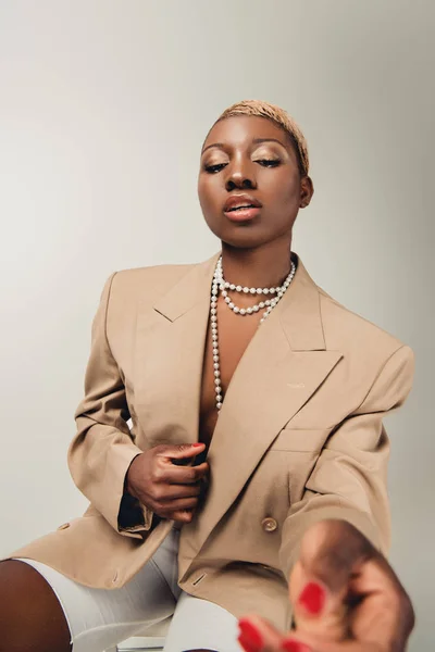 Donna afroamericana alla moda in collana e giacca beige seduta su sedia isolata su grigio — Foto stock