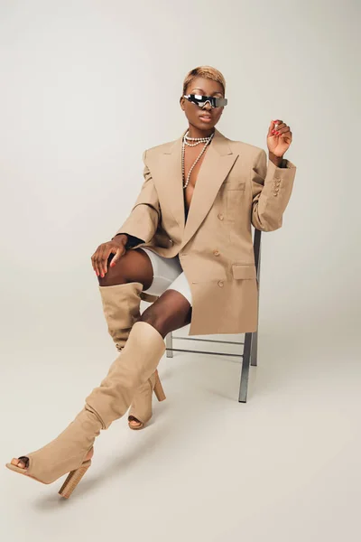 Привлекательная африканская американка в солнцезащитных очках и бежевой куртке, сидящая на стуле на сером — стоковое фото