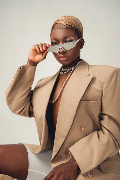 Atractiva chica afroamericana de moda posando en gafas de sol y chaqueta beige aislado en gris - foto de stock
