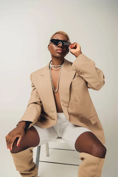 Афроамериканець жінка в сонцезахисні окуляри і модні куртки сидять на стільці на сірий — стокове фото