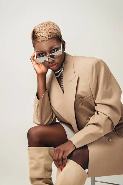 Femme afro-américaine à la mode en lunettes de soleil et veste beige assis sur chaise sur gris — Photo de stock