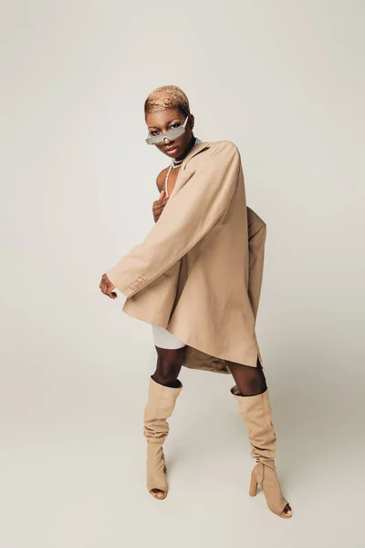 Красивая модная африканская американка, позирующая в солнечных очках и бежевой куртке на сером — стоковое фото
