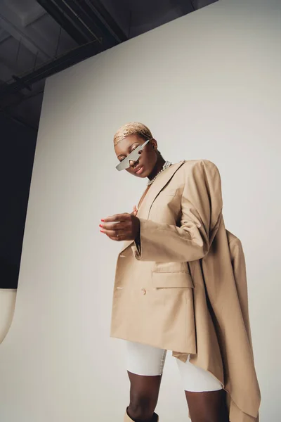 Ragazza afro-americana alla moda in posa con occhiali da sole e giacca beige su grigio — Foto stock