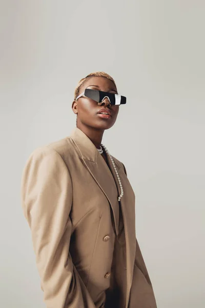 Fashionable african american girl posing in sunglasses and beige jacket isolated on grey — Stock Photo