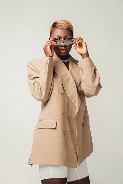 Стильная африканская американская девушка в солнечных очках и элегантном бежевом пиджаке, изолированном на сером — стоковое фото