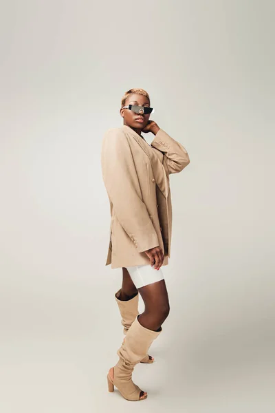 Fashionable african american girl posing in sunglasses and beige jacket on grey — Stock Photo