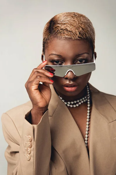 Портрет стильной африканской девушки в солнечных очках и бежевой куртке, изолированной на сером — стоковое фото