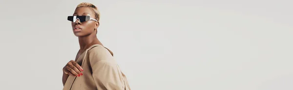 Красивая африканская американка, позирующая в солнечных очках и модной бежевой куртке, изолированной на сером — стоковое фото