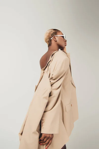 African american girl posing in sunglasses and fashionable beige jacket isolated on grey — Stock Photo