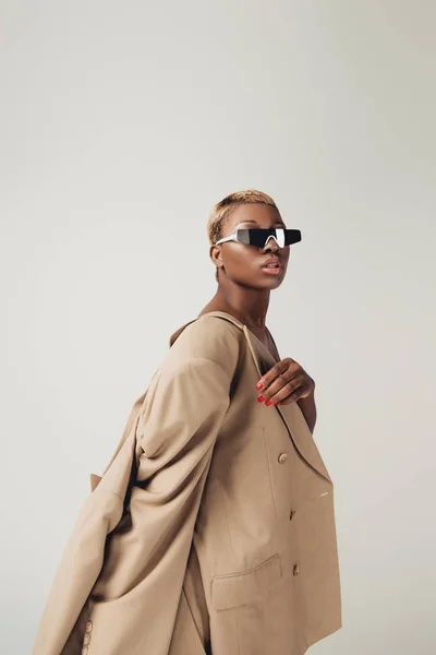 Beautiful stylish african american girl posing in sunglasses and beige jacket isolated on grey — Stock Photo