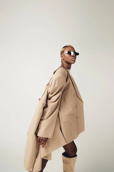 Привлекательная африканская американка, позирующая в солнечных очках и бежевой куртке, изолированная на сером — стоковое фото