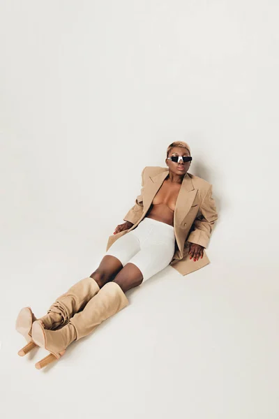 Модная африканская американская девушка позирует в модных солнцезащитных очках и бежевой куртке на сером — стоковое фото