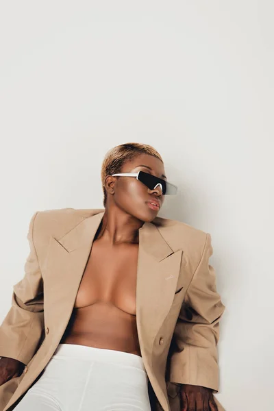 Chica afroamericana elegante posando en gafas de sol y chaqueta beige aislado en gris - foto de stock