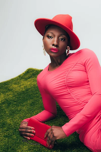 Mujer afroamericana de moda en vestido rosa y sombrero posando con bolsa sobre hierba verde - foto de stock